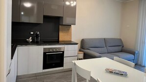 Een moderne keuken met donkere kastjes, een zwarte kookplaat en een grijze bank in de woonkamer.