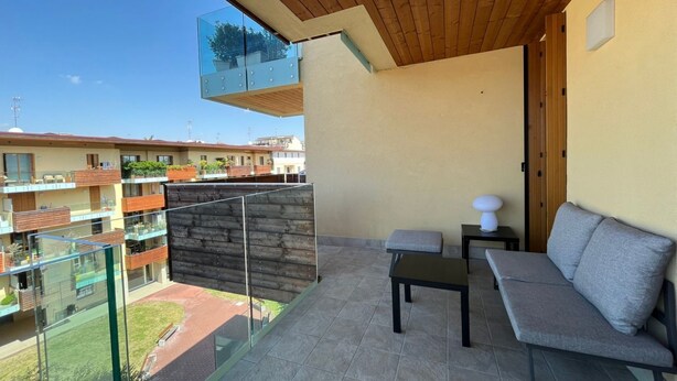 Een balkon met een glazen hekwerk, een houten plafond, een grijze bank en een kleine tafel.