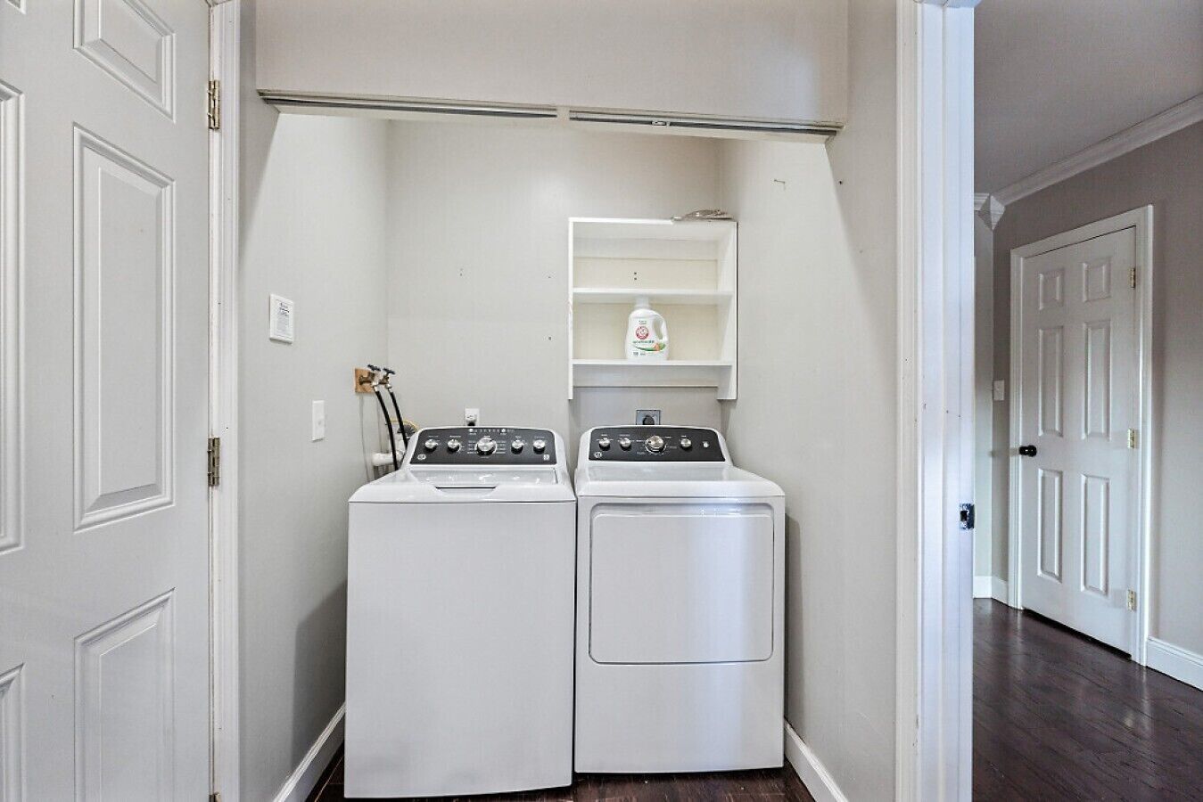 A laundry room with two white front-loading washers and dryers, a metal shelf above the dryers, and a white door on the left.