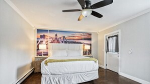 Una camera da letto con un letto, un ventilatore a soffitto, un comodino, una lampada, una porta e una parete con un dipinto di un tramonto.