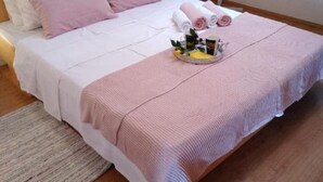 A neatly made bed with pink bedding, a bedside table with a lamp, and a tray with towels and a flower arrangement.