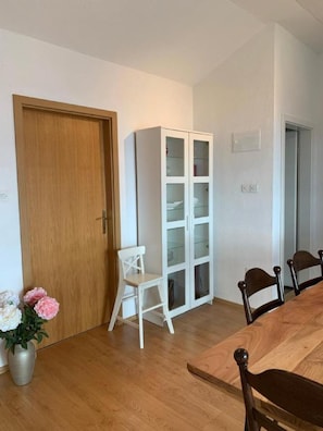 Una habitación con puerta de madera, un armario blanco con puertas de cristal, una mesa de comedor con sillas y una planta en maceta.