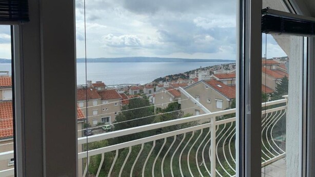 A view from a balcony with a railing, overlooking a residential area with red-tiled roofs and a body of water in the distance.
