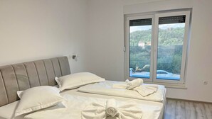 Un lit bien fait, avec des oreillers et des serviettes, une fenêtre donnant sur un paysage verdoyant et un véhicule, et un plancher en bois de couleur claire.