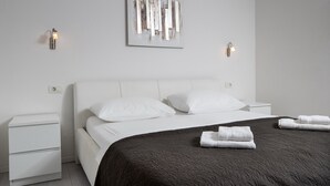 A modern bedroom with a white bed, two white pillows, a dark quilted bedspread, two bedside tables, and two wall-mounted light fixtures.