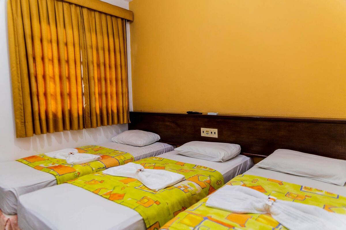 A hotel room with three single beds, each with a yellow and orange patterned bedspread, white pillows, and neatly folded white towels. The room has a wooden headboard, a window with vertical blinds, and a wall socket.