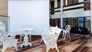 Amplio espacio interior con muebles blancos ornamentados, incluyendo sillas y una mesa, frente a un edificio de varios niveles con grandes ventanales y vigas de madera.