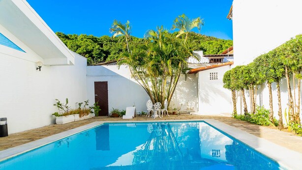Una piscina con un muro blanco y una puerta, rodeada de follaje y un cielo azul despejado.