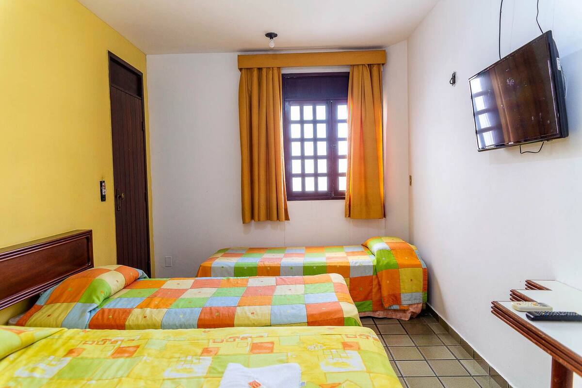 A room with two beds, a window with curtains, a television on the wall, and a desk with a remote control.