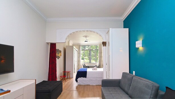 Ein modernes Wohnzimmer mit grauem Sofa, einem schwarzen Hocker, einem Flachbildfernseher und einem Bett mit blauer Decke.