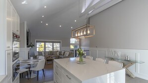 A modern kitchen with white cabinetry, a central island, and a dining area with a glass table. - Chic Hamptons Home near beach! (Hampton Bays)