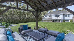 A covered patio area with outdoor furniture, a lawn, and a house in the background. - Chic Hamptons Home near beach! (Hampton Bays)