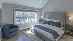 A bedroom with a large bed, a blue armchair, a window with a view, and a ceiling light. - Chic Hamptons Home near beach! (Hampton Bays)