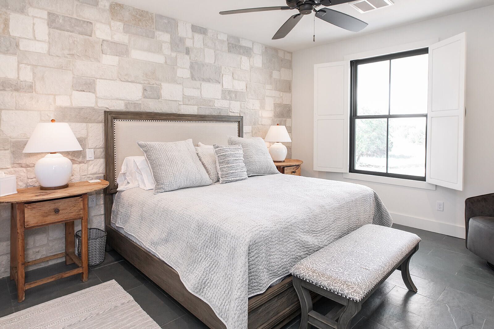 A bedroom with a stone wall, a bed with a headboard, two bedside tables with lamps, a bench, and a window with blinds.