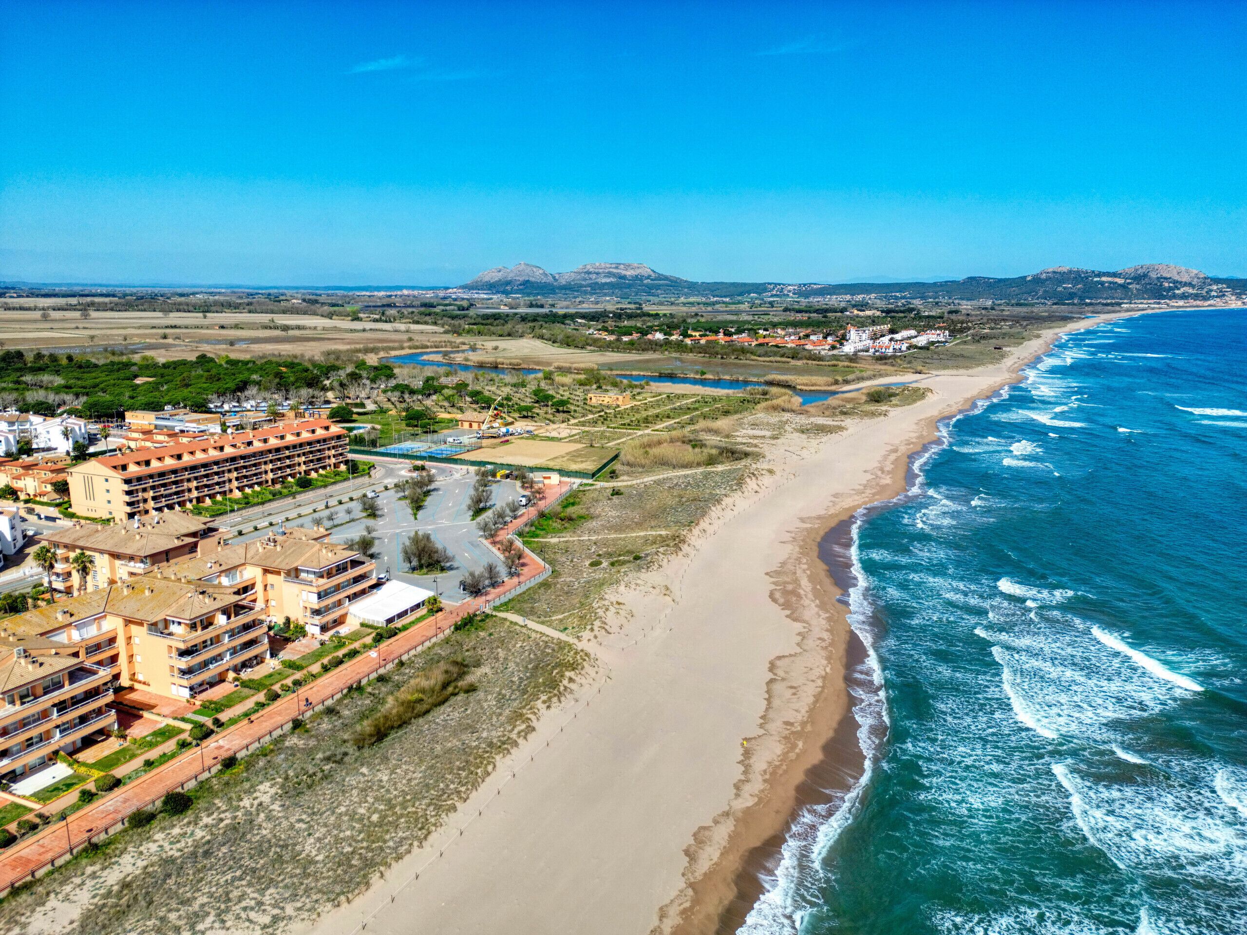 An aerial view of a coastal resort with beachfront properties, a marina, and a mountainous backdrop.