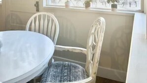 Un coin repas avec une table ronde et deux chaises blanches, un lustre et une fenêtre donnant sur des arbres et une clôture.