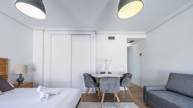 A modern hotel room with a bed, a grey sofa, a dining table with chairs, and a small table with a vase. - Ático del Oso · Terraza privada en Lavapiés (Madrid)