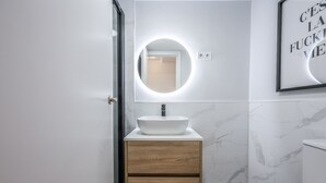 A modern bathroom with a round mirror, a white sink, and a wooden vanity. - Ático del Oso · Terraza privada en Lavapiés (Madrid)