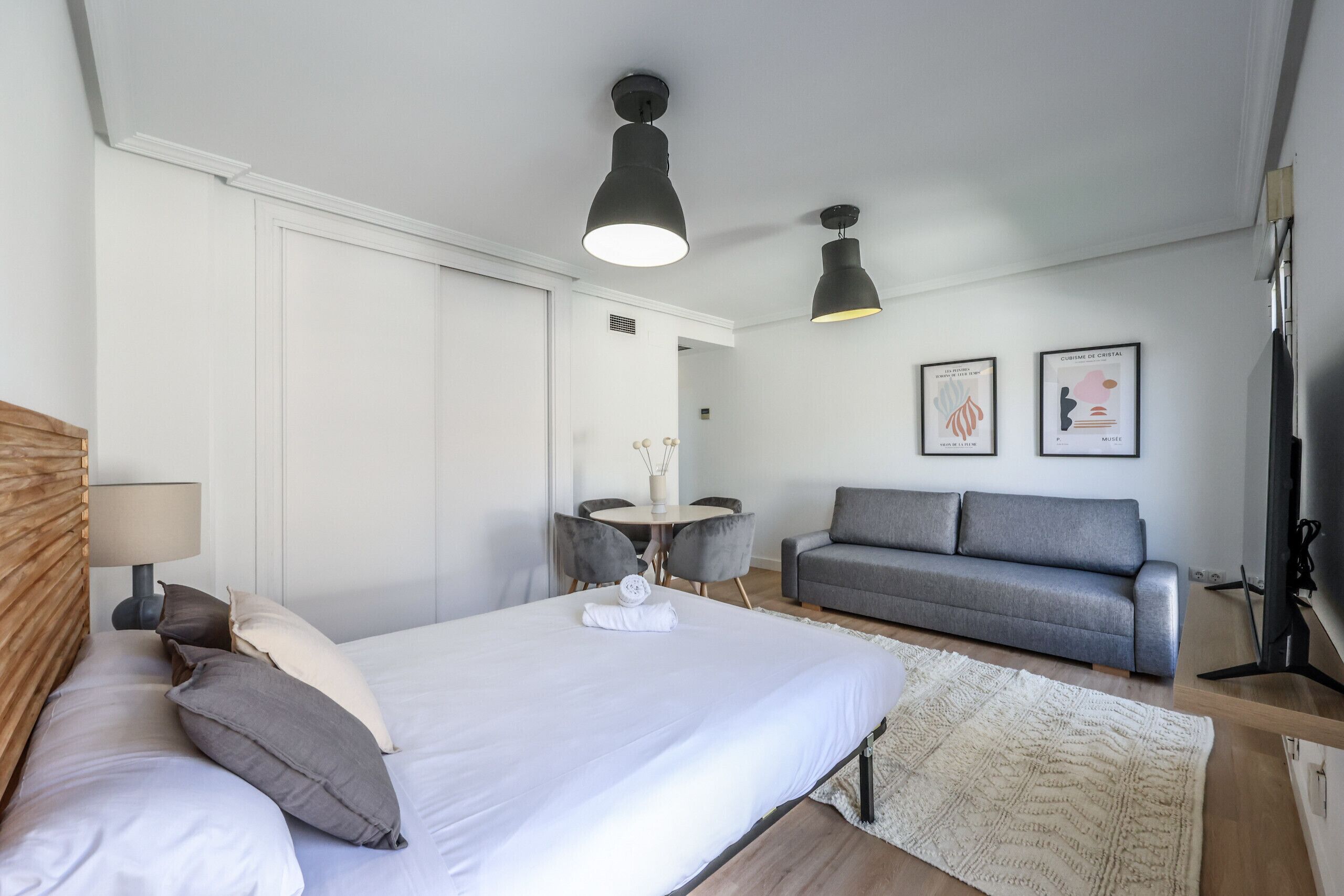 A modern bedroom with a large bed, a grey sofa, a small round table with chairs, and two pendant lights.