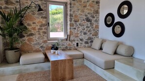 A cozy living room with a stone wall, a wooden coffee table, a beige sofa, and a large window. - Stone build Suite with private small pool (Skiathos)