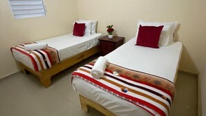 A small, clean room with two single beds, each with striped bedding and red pillows. There is a small table with a vase of flowers between the beds.