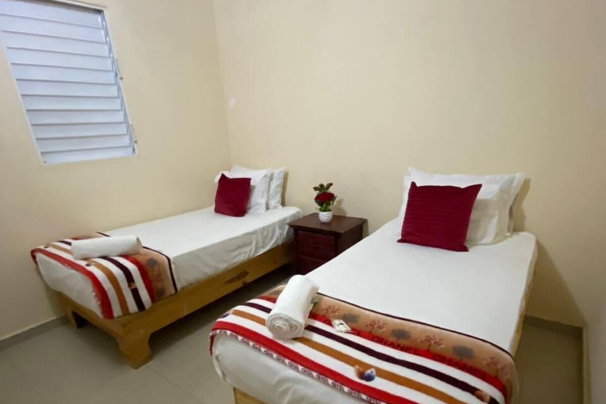 A small, clean room with two single beds, each with striped bedding and red pillows. There is a small table with a vase of flowers between the beds.