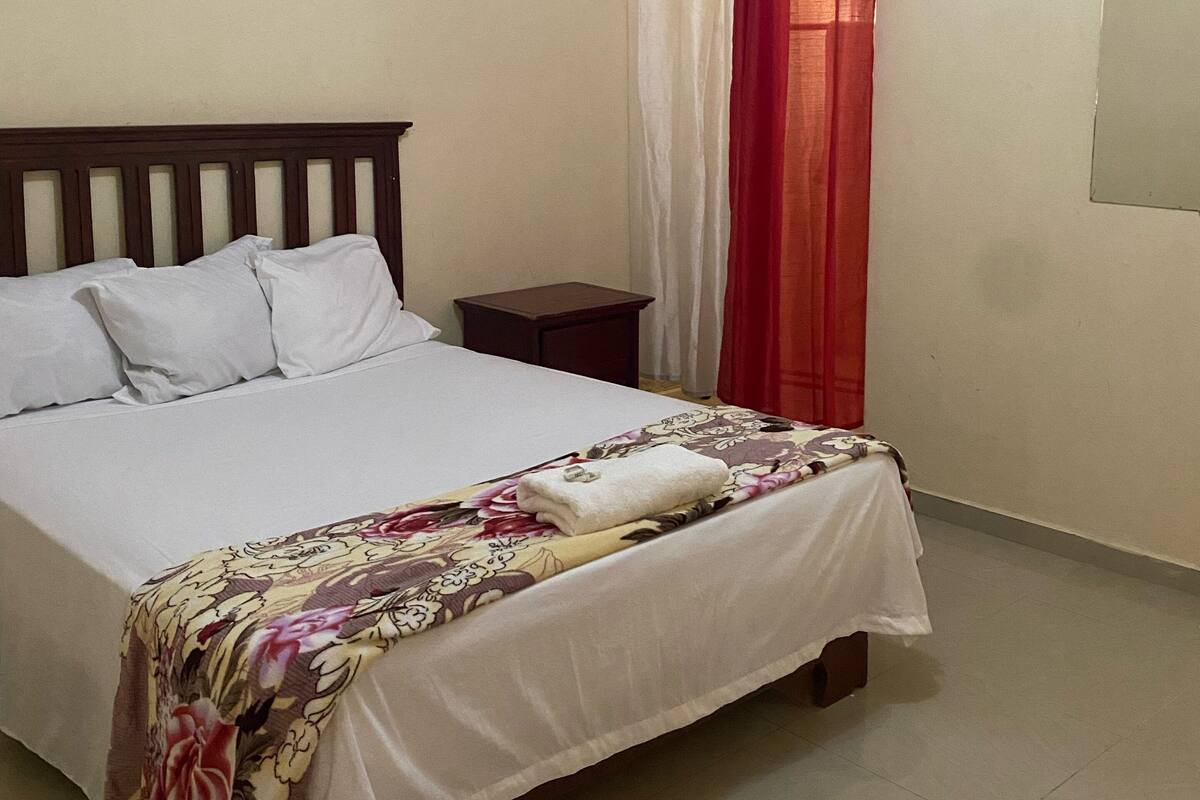 A neatly made bed with a floral bedspread and white pillows, a small bedside table, and a window with red curtains.