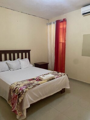 A neatly made bed with a floral bedspread and white pillows, a small bedside table, and a window with red curtains. - Hotel Plaza la Duarte (San Juan de la Maguana)