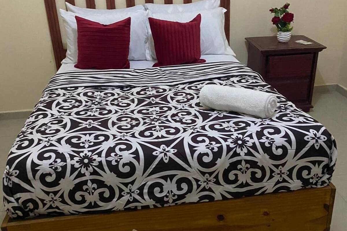 A bed with a patterned bedspread, two red pillows, and a white towel. There is a small wooden nightstand with a vase of flowers to the right.