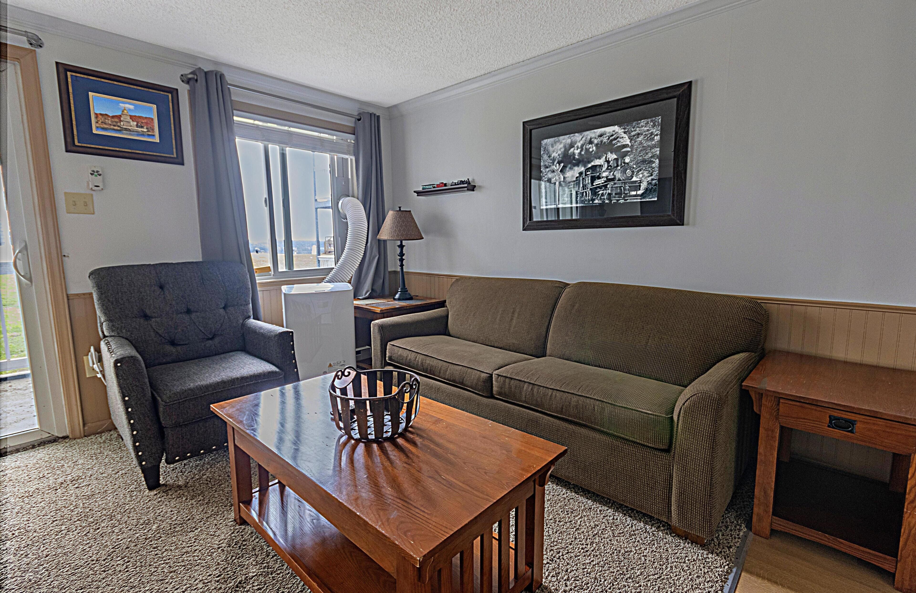 A living room with a grey armchair, a brown sofa, a wooden coffee table, and a lamp. There are two framed pictures on the wall and a window with curtains.