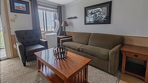En stue med en grå lænestol, en brun sofa, et træsofabord og en lampe. Der er to billeder i rammer på væggen og et vindue med gardiner.