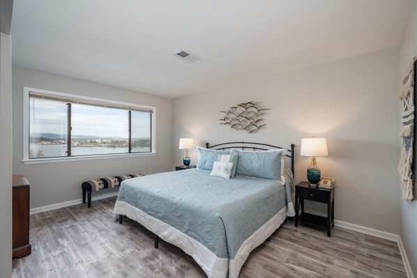 A bedroom with a large window, a bed with a blue bedspread, two bedside tables with lamps, and a decorative wall piece.