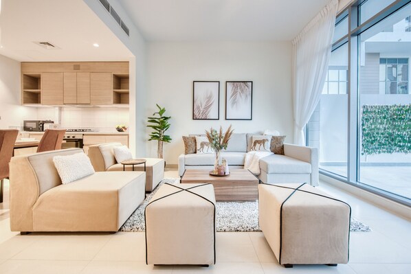 A modern living room with a sofa set, a coffee table, and a dining area with chairs. There is a kitchen in the background with wooden cabinets and a microwave.