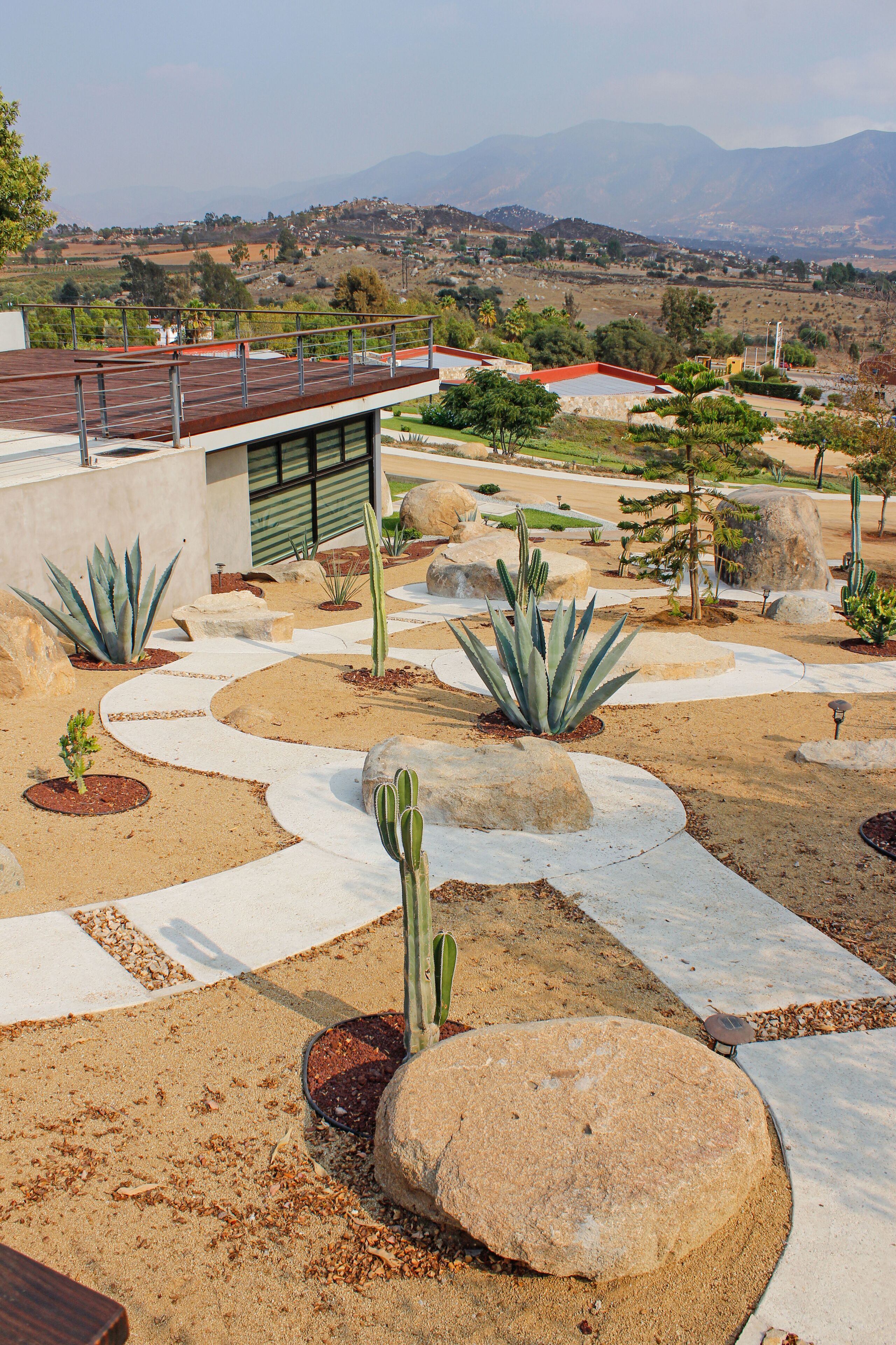 Um jardim com cactos e pedras, um edifício com telhado vermelho e vista para colinas distantes.