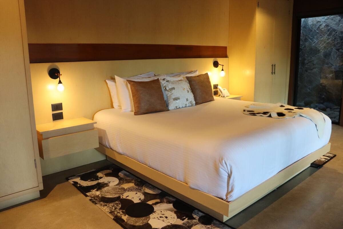 A modern bedroom with a large bed, bedside lamps, a nightstand, and a view of a stone wall through a window.
