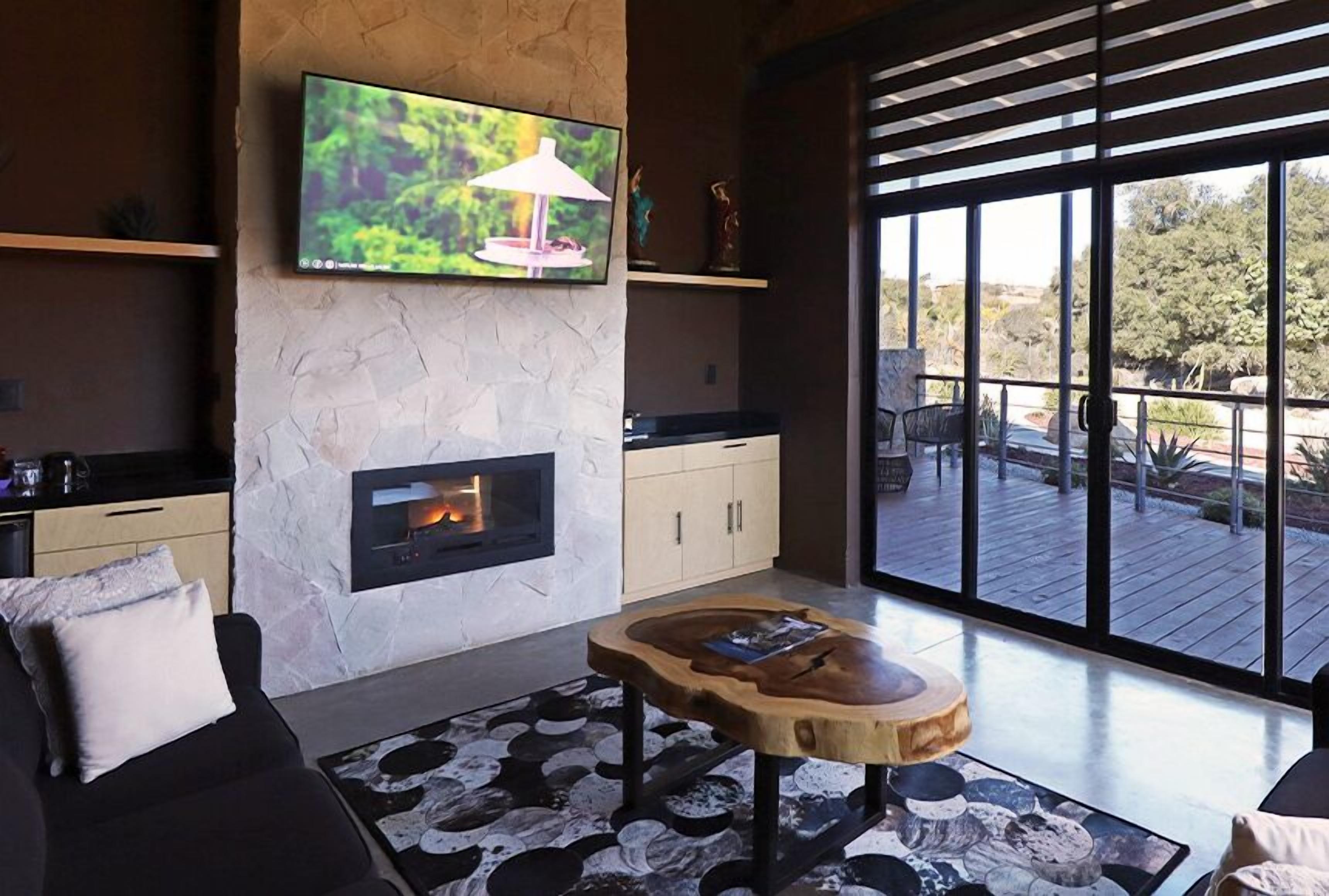 A modern living room with a stone fireplace, a flat-screen TV mounted above it, and a large window offering a view of the outdoors.