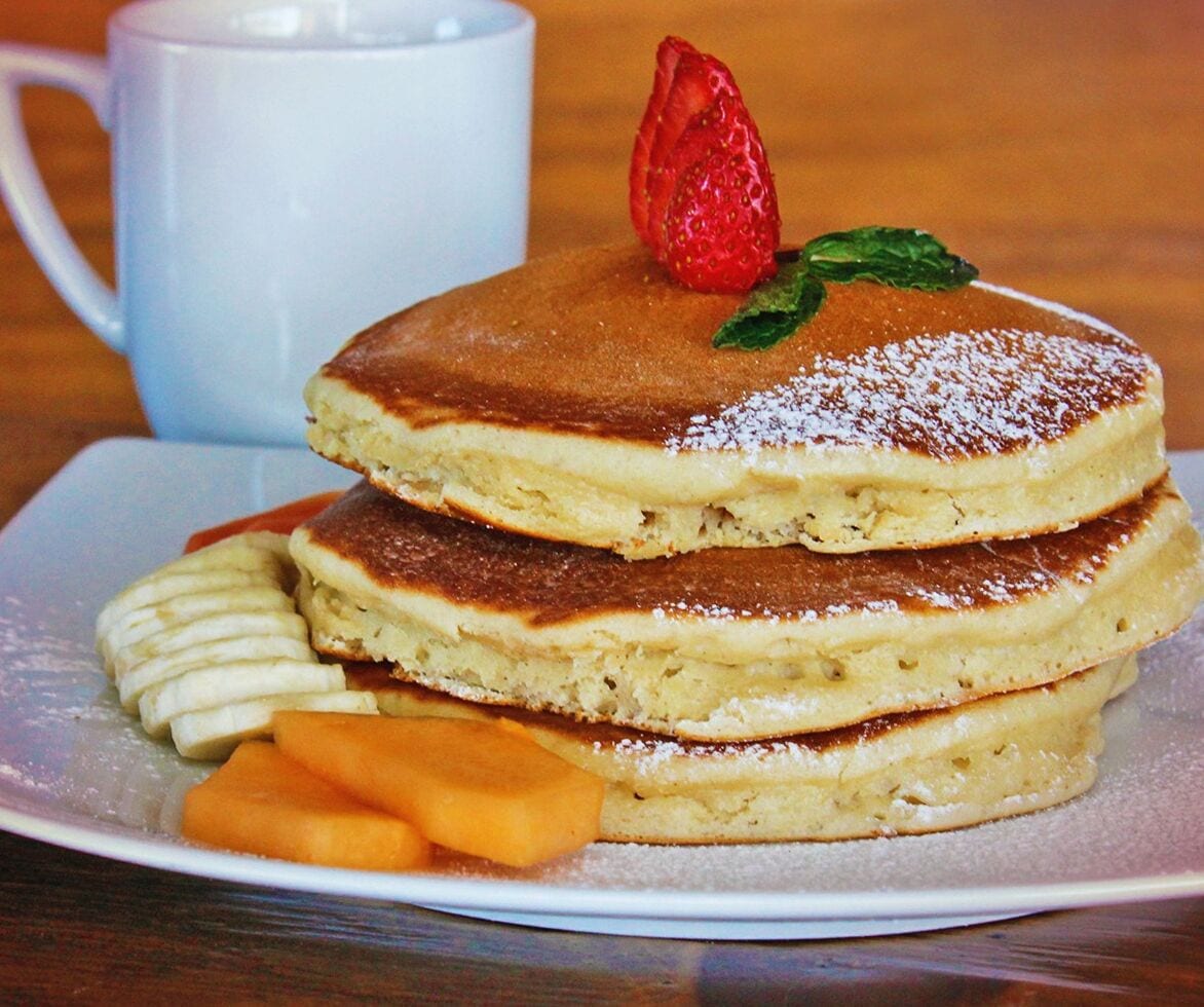 Una pila de tortitas coronadas con fresa y menta, acompañadas de rodajas de plátano y una taza de café.