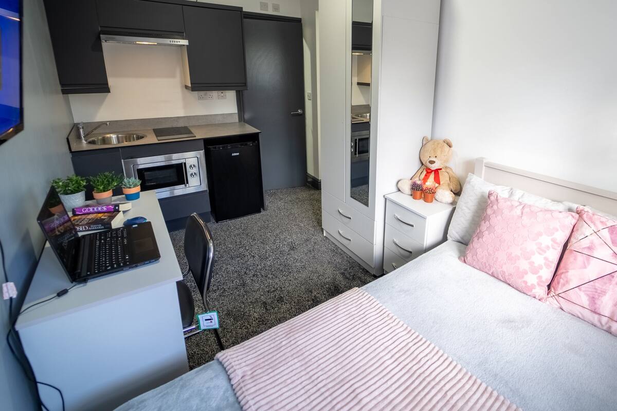 Ein kleines Zimmer mit Küchenzeile, einem Bett mit rosa Kissen und einem Teddy auf dem Nachttisch.