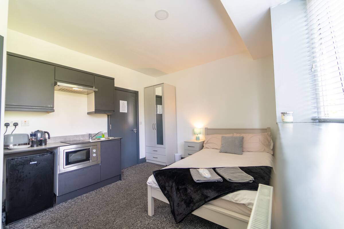 Ein kompaktes Hotelzimmer mit Küchenzeile, einem Bett mit schwarzer Decke und einem Nachttisch mit Lampe.