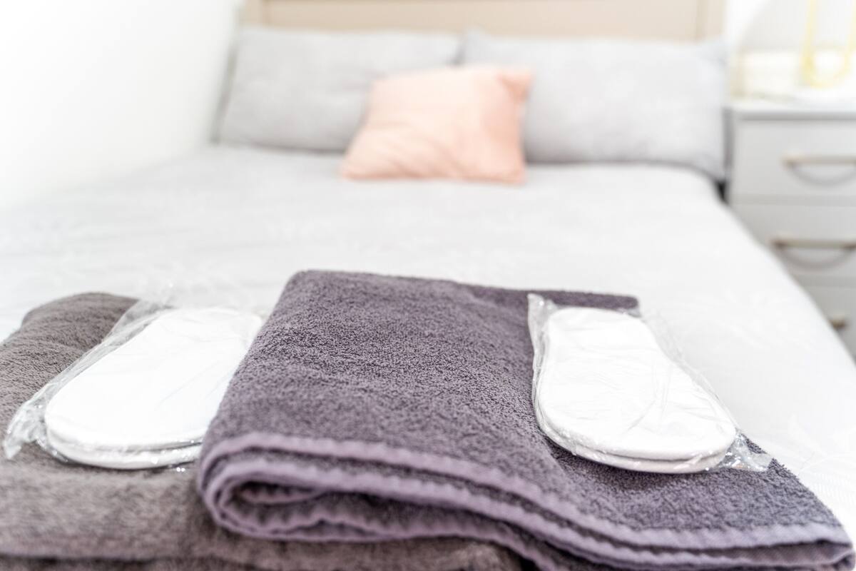 Ein ordentlich bezogenes Bett mit einer grau-weißen Decke, einem gefalteten violetten Handtuch und zwei weißen Hausschuhen.