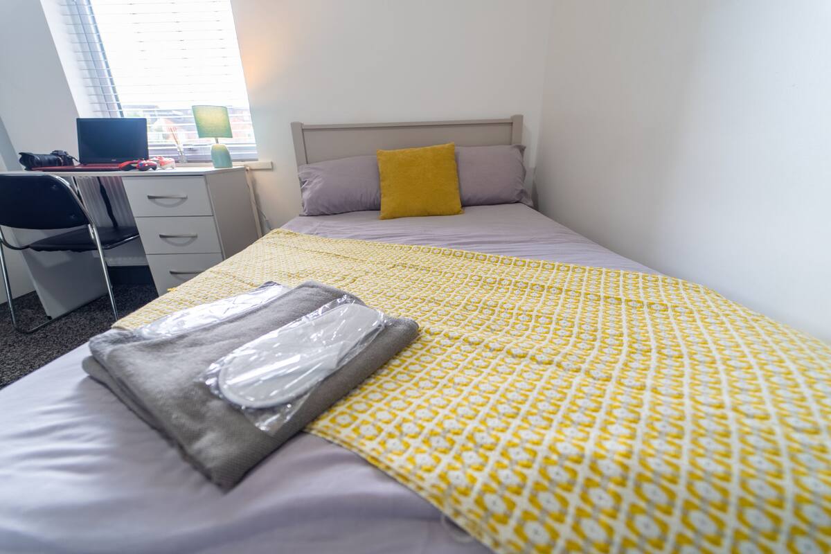 Ein ordentlich gemachtes Bett mit einer gelb gemusterten Decke, einem grauen Kopfkissen und einem senffarbenen Kissen. Links befindet sich ein Schreibtisch mit einem Laptop und einer Lampe.
