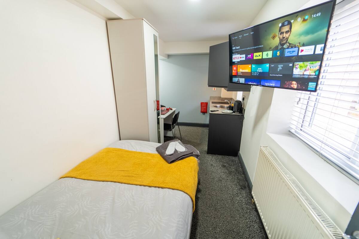 Ein Hotelzimmer mit Bett, Wandfernseher, Schreibtisch mit Stuhl und Fenster mit Jalousien.