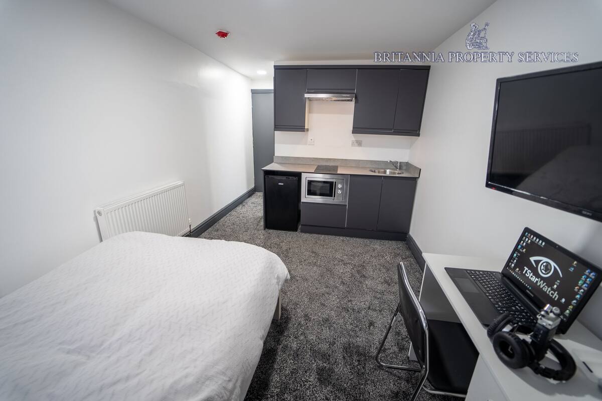 Ein kleines Hotelzimmer mit Bett, Küchenzeile und Schreibtisch, auf dem ein Laptop und ein TStarWatch Gerät liegen.