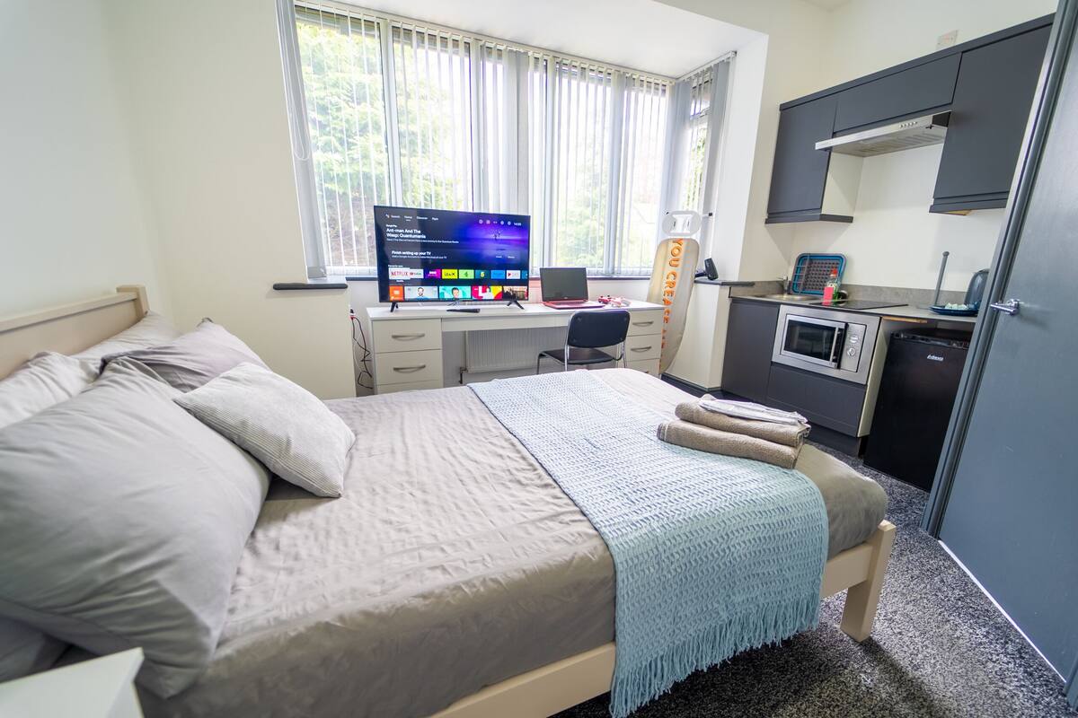 Ein Schlafzimmer mit einem Bett, einem Schreibtisch mit Computer, einer Mikrowelle und einer kleinen Küchenzeile.