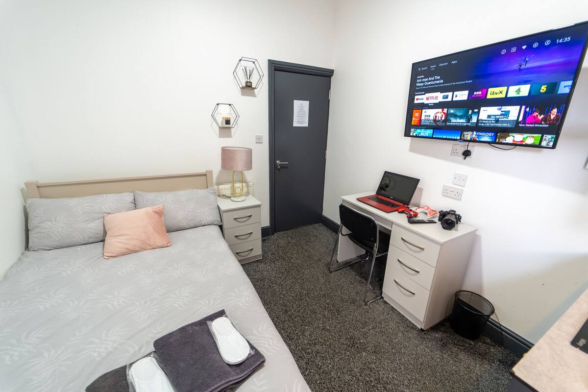 Ein Schlafzimmer mit einem Bett, einem Schreibtisch, einem Computer und einem Fernseher, der verschiedene Sender zeigt.