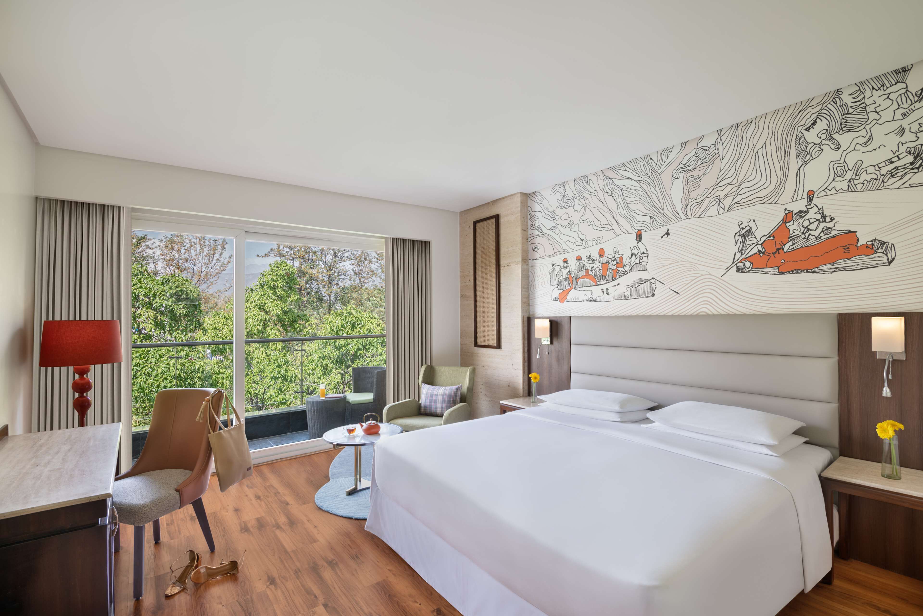 A hotel room with a large bed, a desk, a chair, a small table, and a view of greenery through large windows.