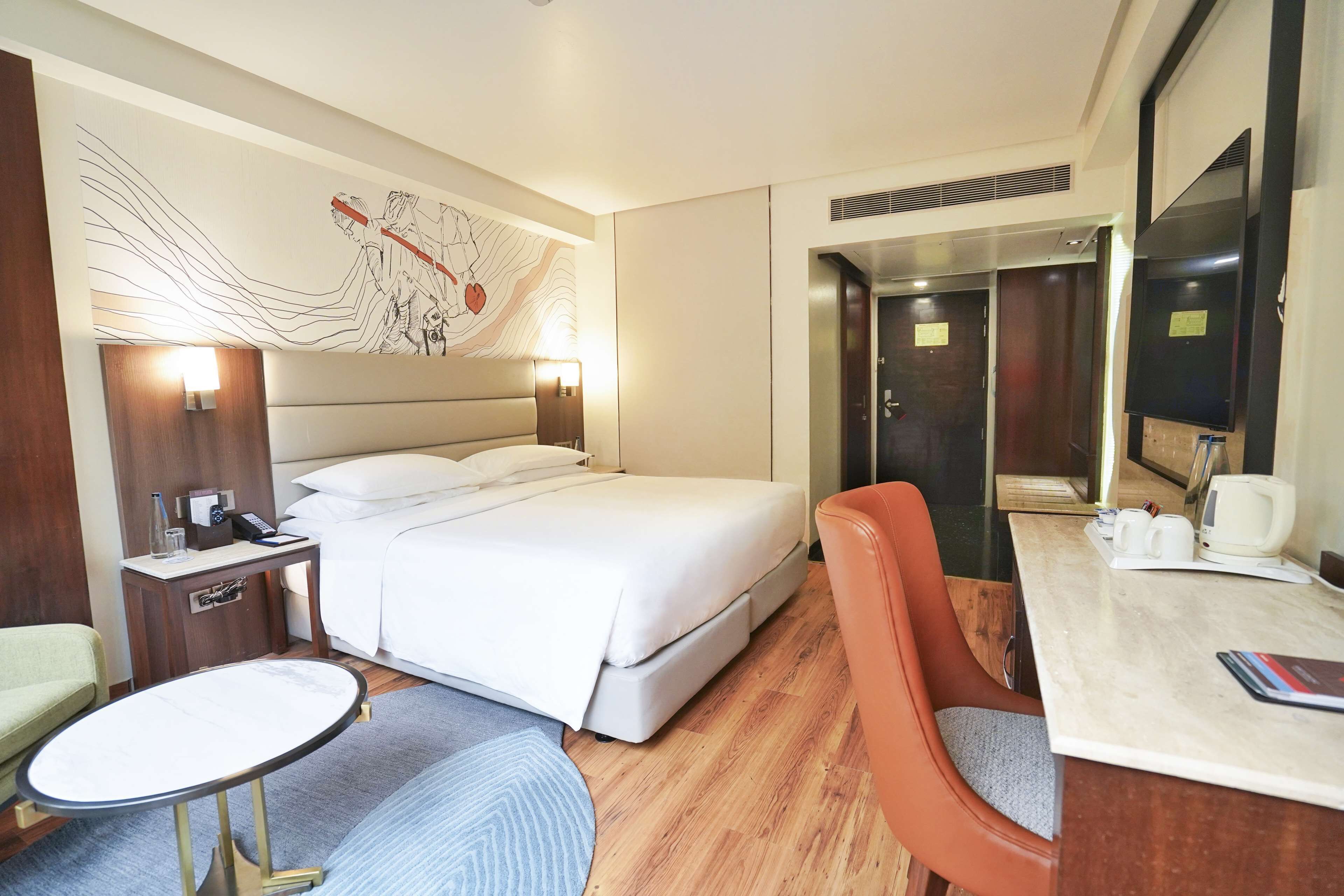 A hotel room with a bed, a desk, a chair, a small table, a wall-mounted artwork, and a mirror.
