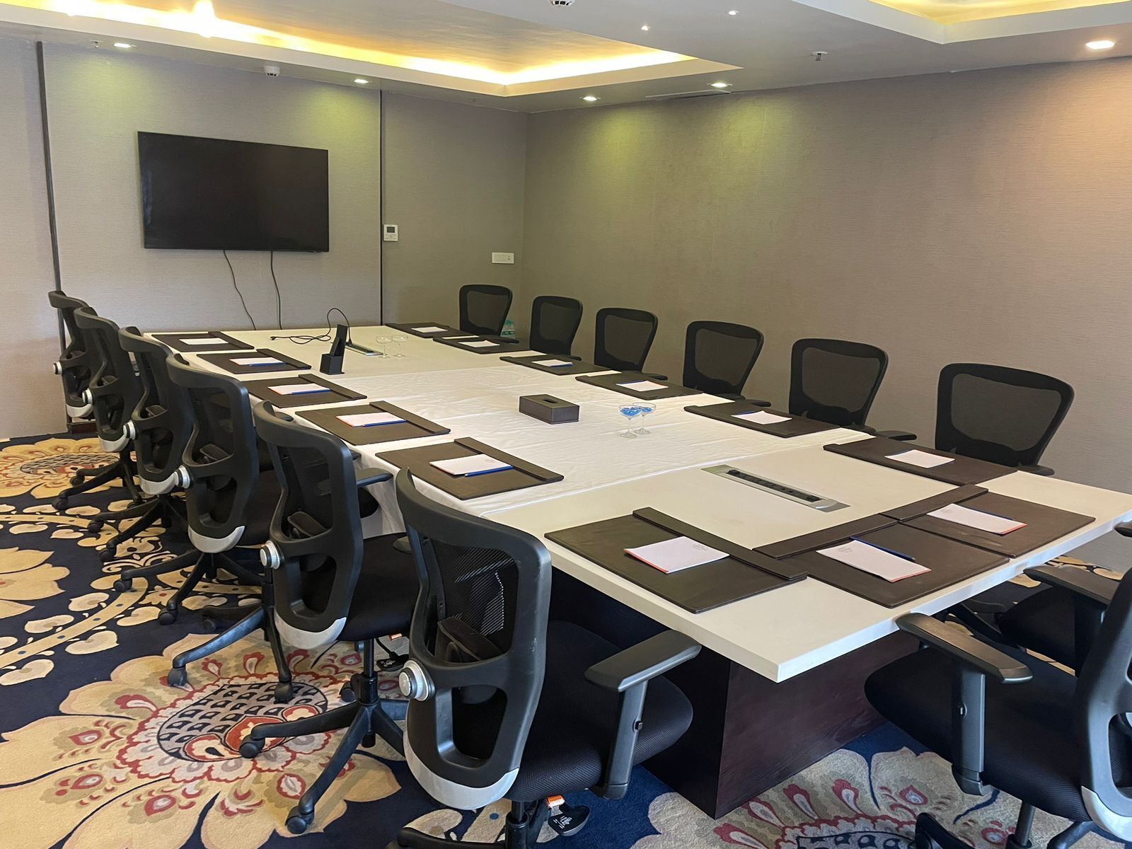 A conference room with a long table, chairs, a flat-screen TV, and a carpeted floor.