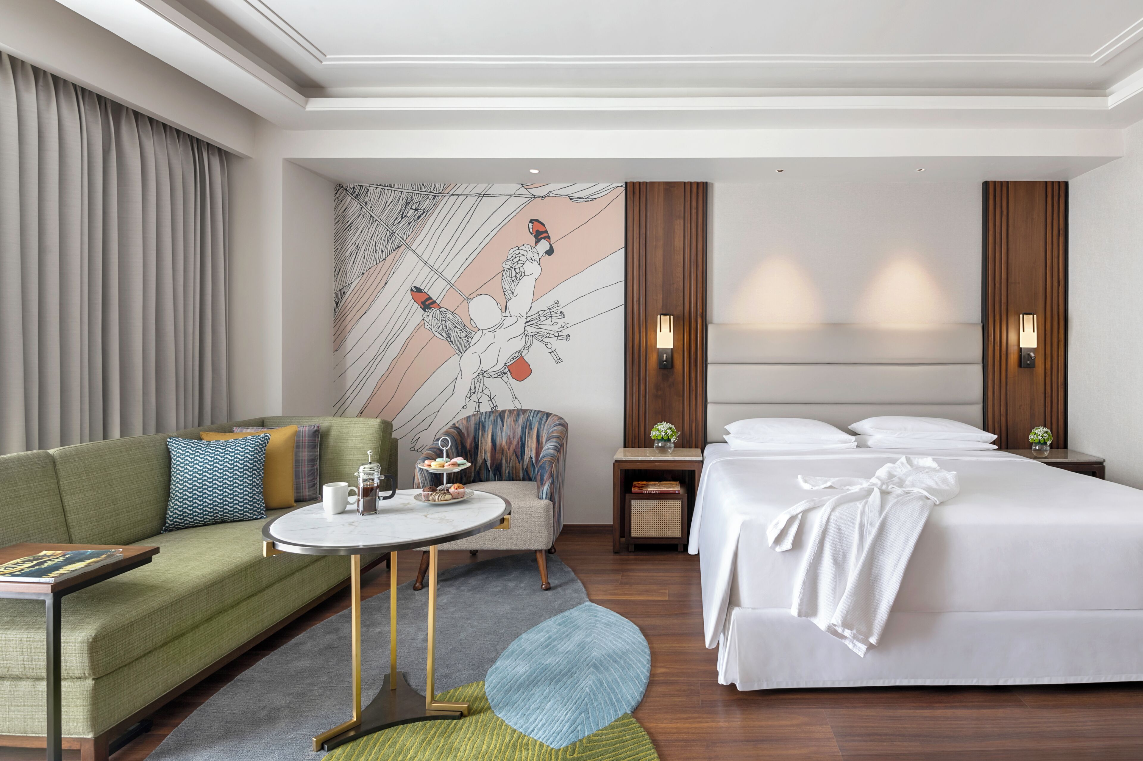 A hotel room with a green sofa, a round coffee table, a bed with white linens, a chair, and a wall mural.