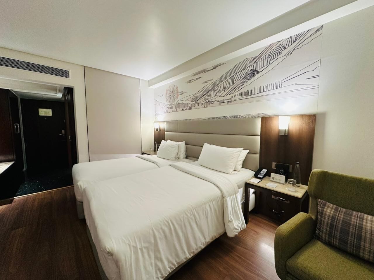 Una habitación de hotel con dos camas, un sillón verde, una mesita de noche con una lámpara y una obra de arte en la pared.